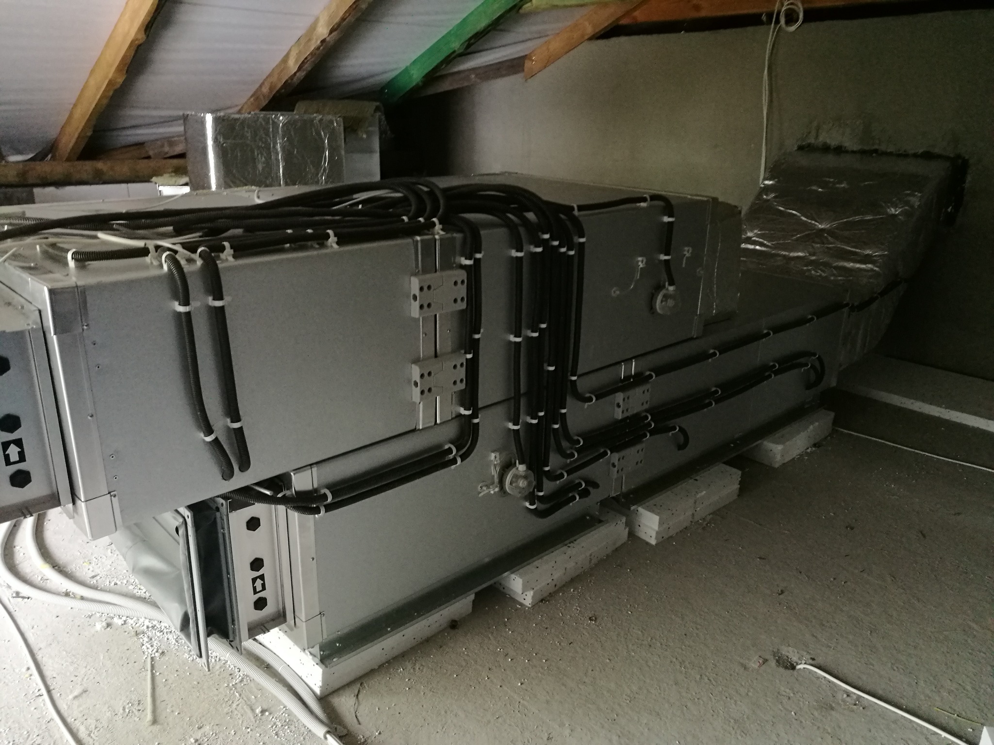 Szara centrala wentylacyjna z licznymi przewodami w surowym pomieszczeniu na poddaszu, widoczne elementy konstrukcji dachu.