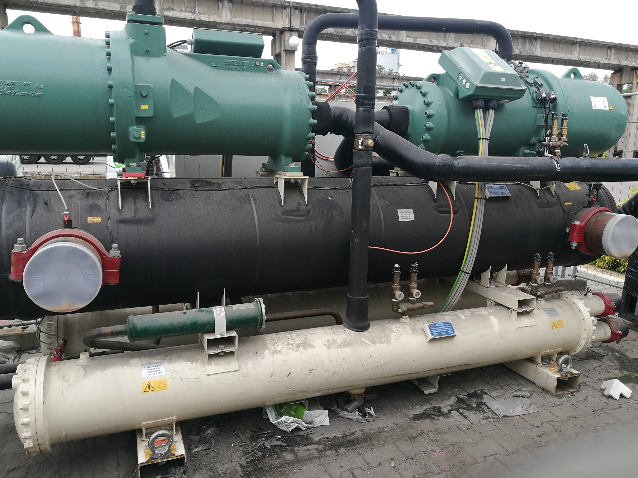 Złożony układ chłodniczy z rurami i zaworami, z elementami w kolorze zielonym, czarnym i kremowym, umieszczony na zewnątrz budynku na betonowej powierzchni.