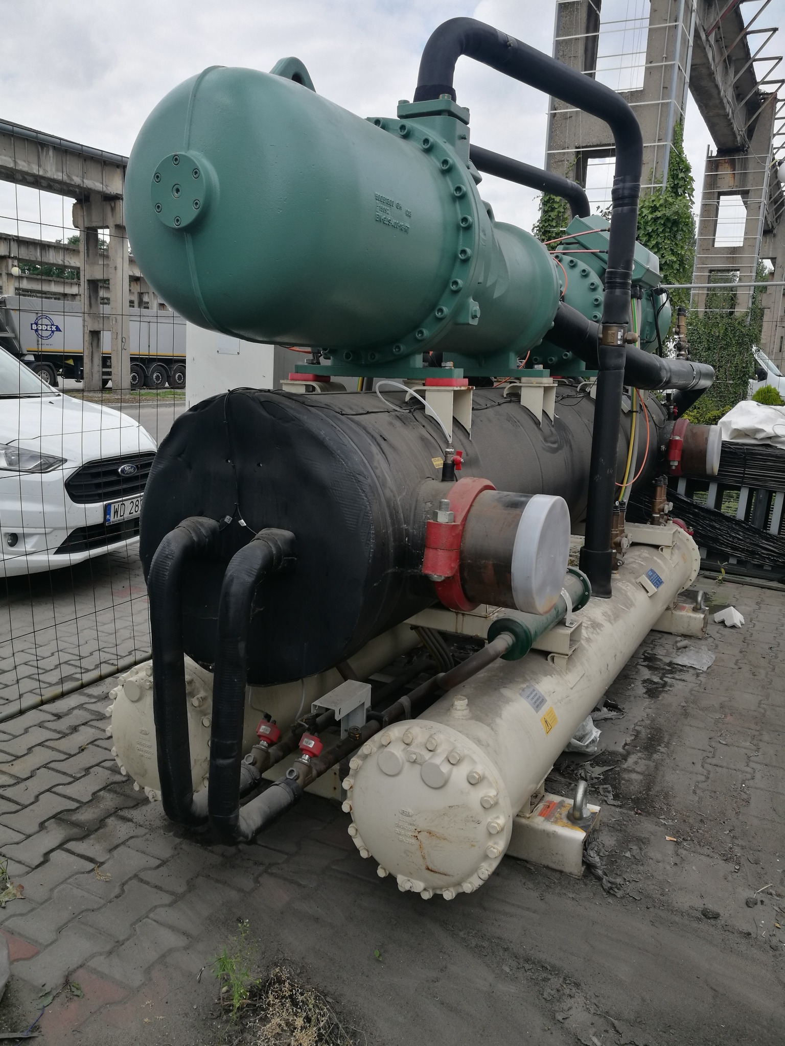 Duży agregat chłodniczy w kolorze zielono-czarnym, z widocznymi rurami i elementami instalacji, na zewnątrz budynku przemysłowego, częściowo zasłonięty siatką ogrodzeniową.