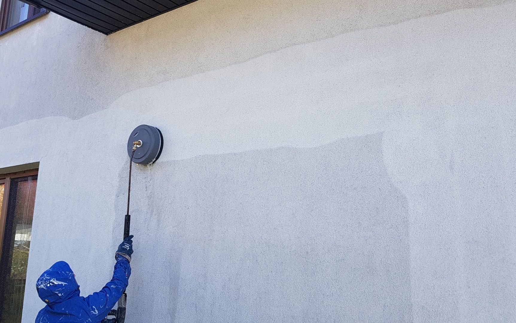 Pracownik w niebieskim kombinezonie czyści elewację budynku za pomocą myjki ciśnieniowej z okrągłą dyszą, widoczne różnice w odcieniu elewacji przed i po czyszczeniu.