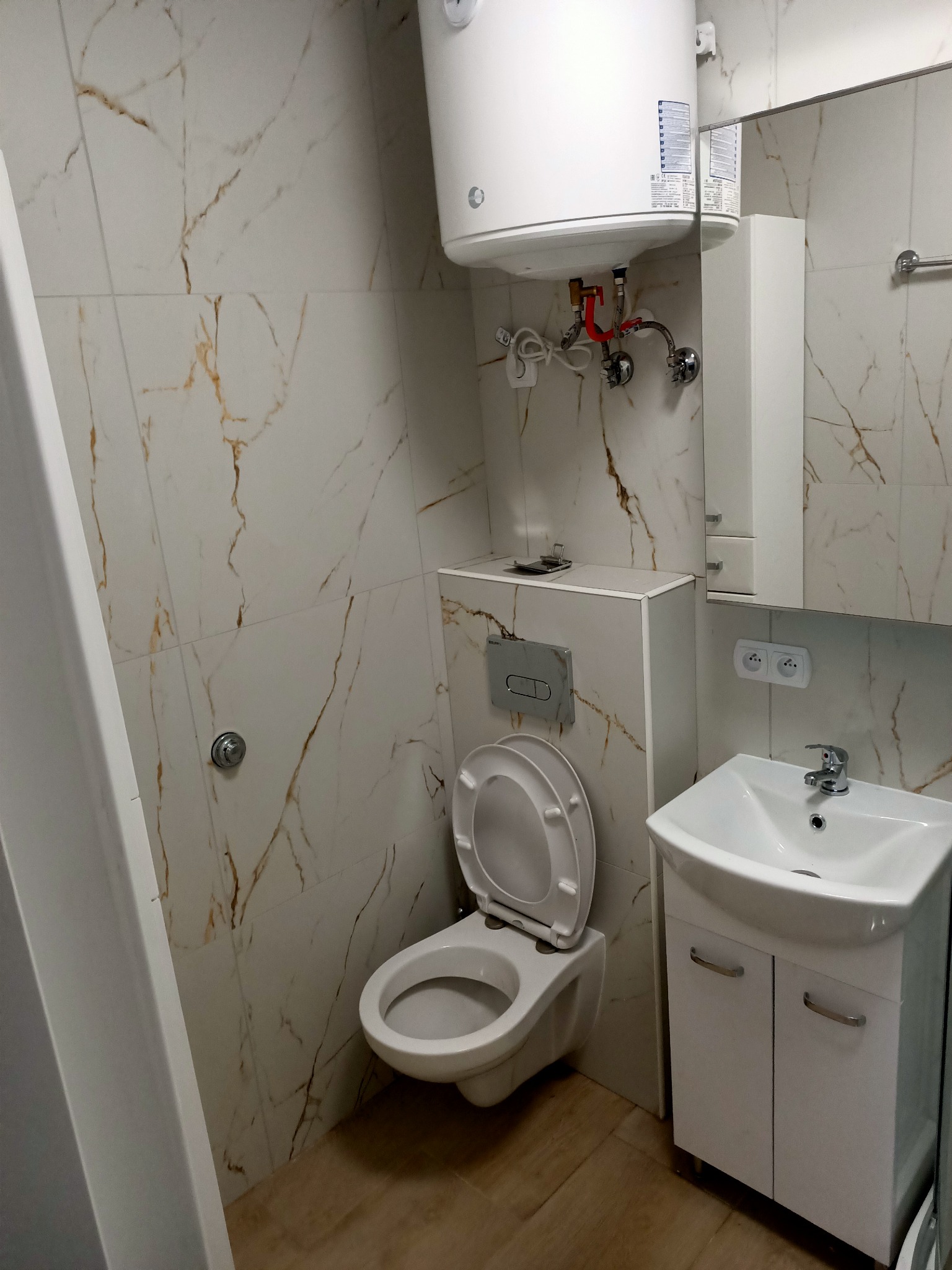 Nowoczesna łazienka z marmurowymi płytkami ściennymi, podwieszaną toaletą z otwartą klapą, białą szafką pod umywalką i bojlerem elektrycznym nad zabudową toalety.