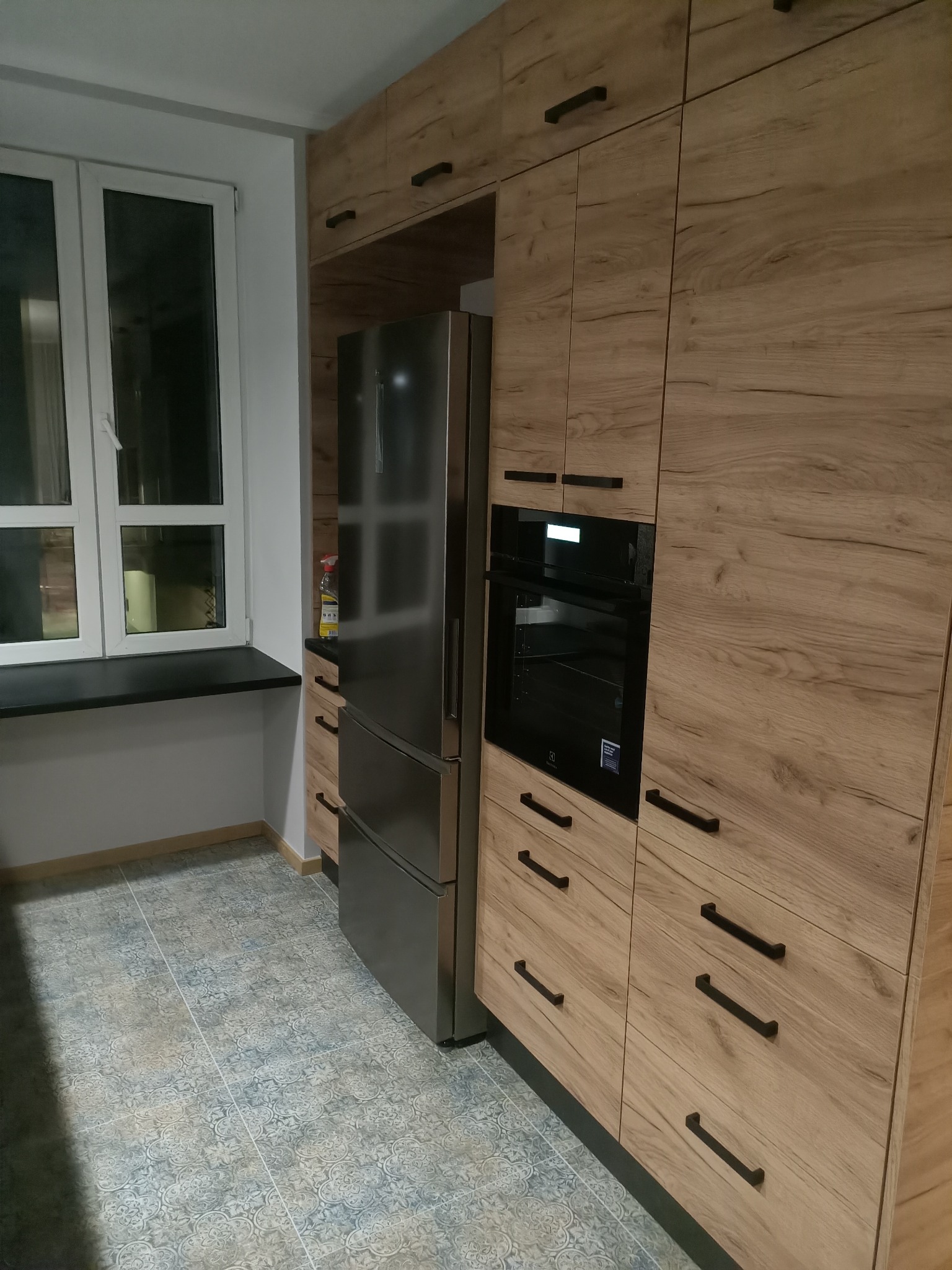 Nowoczesna kuchnia z drewnianymi szafkami, zabudowanym piekarnikiem Electrolux, lodówką side-by-side i wzorzystą podłogą z płytek ceramicznych.