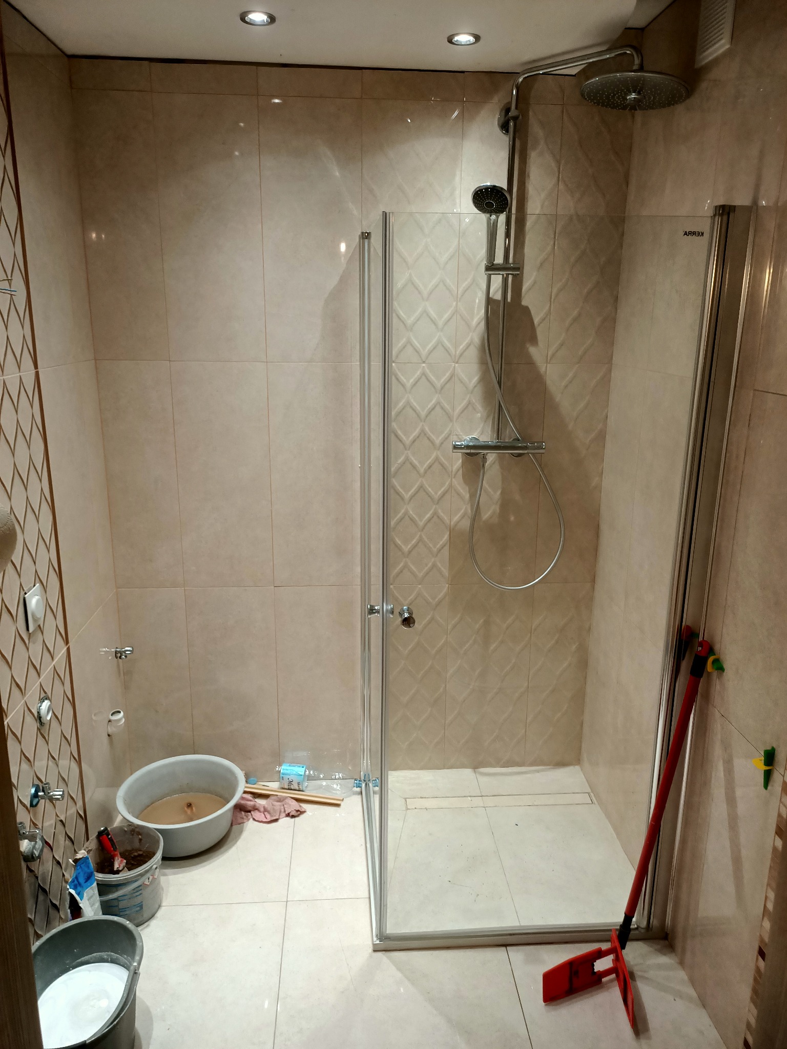 Łazienka w trakcie remontu: widoczna kabina prysznicowa z deszczownicą, wiadro z zaprawą, mop oparty o ścianę, płytki ceramiczne na ścianach.
