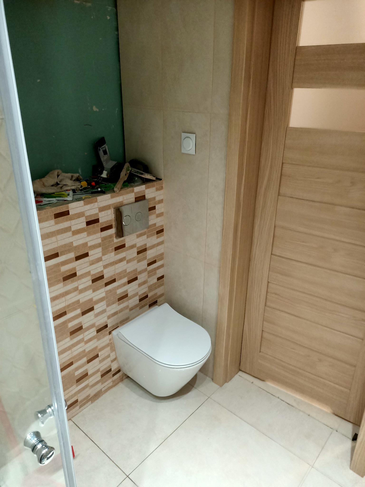 Ściana z mozaiką w łazience, z zamontowaną podtynkową spłuczką i wiszącą toaletą, widoczne narzędzia na półce, drzwi z jasnego drewna z przeszkleniami.