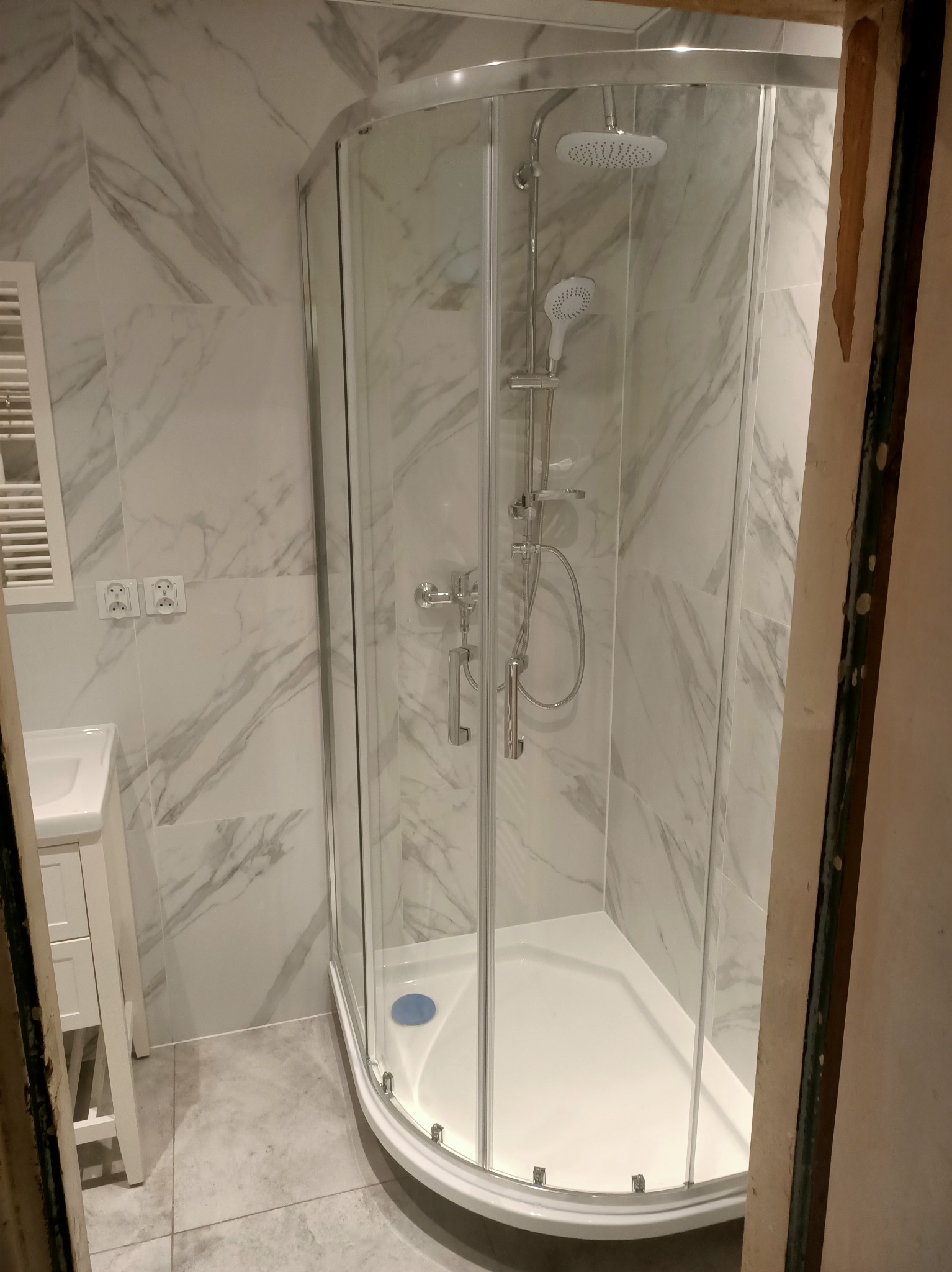 Narożna kabina prysznicowa z hydromasażem i deszczownicą, marmurowe płytki ścienne, biała umywalka z szafką, widoczne fragmenty ościeżnicy w trakcie remontu.