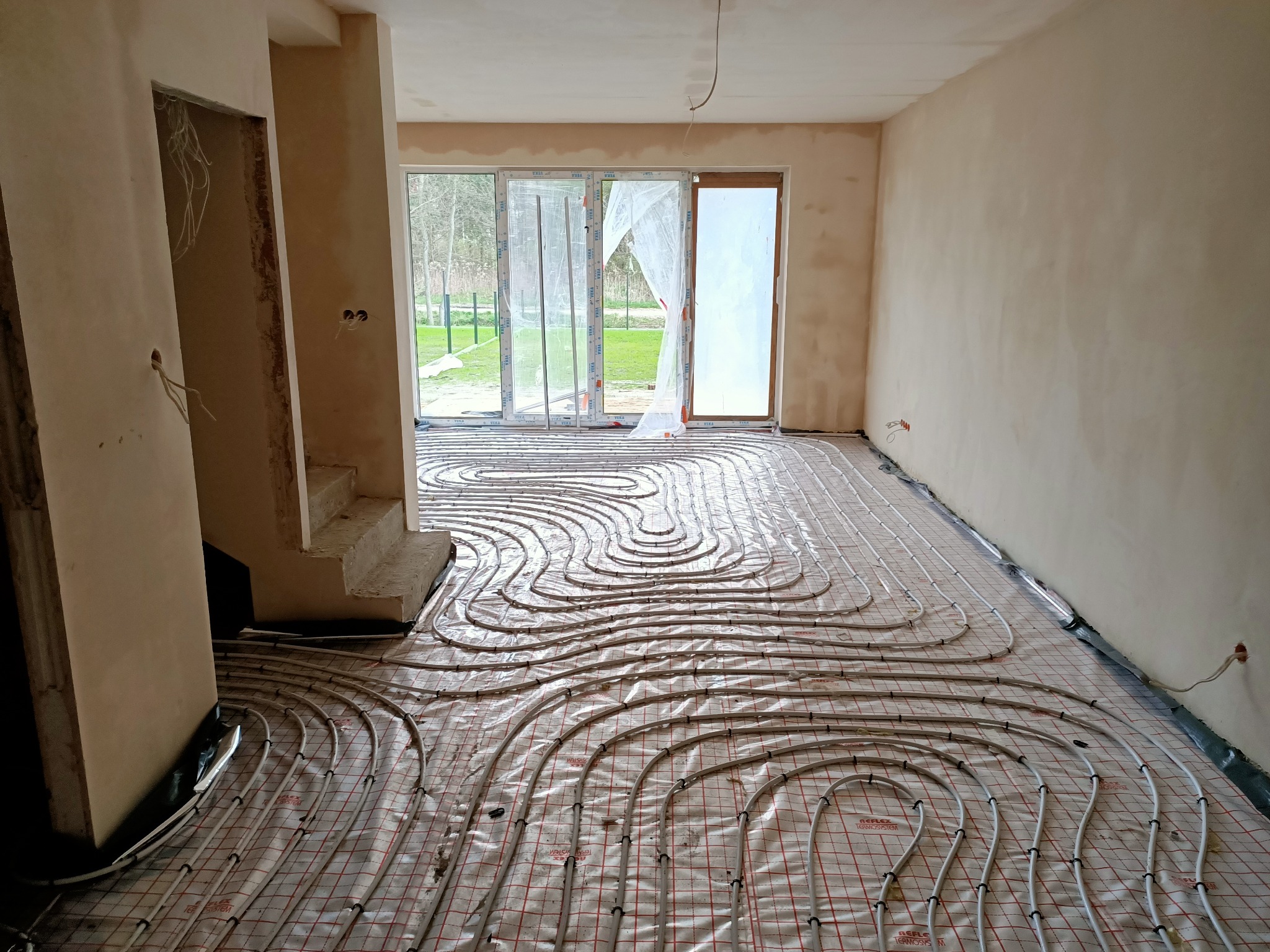 Rury systemu ogrzewania podłogowego ułożone na folii z siatką w surowym wnętrzu przed wylewką, widoczne okna balkonowe i przygotowane ściany.