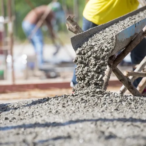 Świeży beton wylewany z metalowej rynny na drewnianym stelażu, z pracownikami w tle. Jasne, naturalne światło podkreśla fakturę betonu.