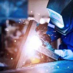 Spawacz w niebieskim kombinezonie i masce spawalniczej łączy metalowe profile, iskry rozbryzgują się wokół miejsca spawania.