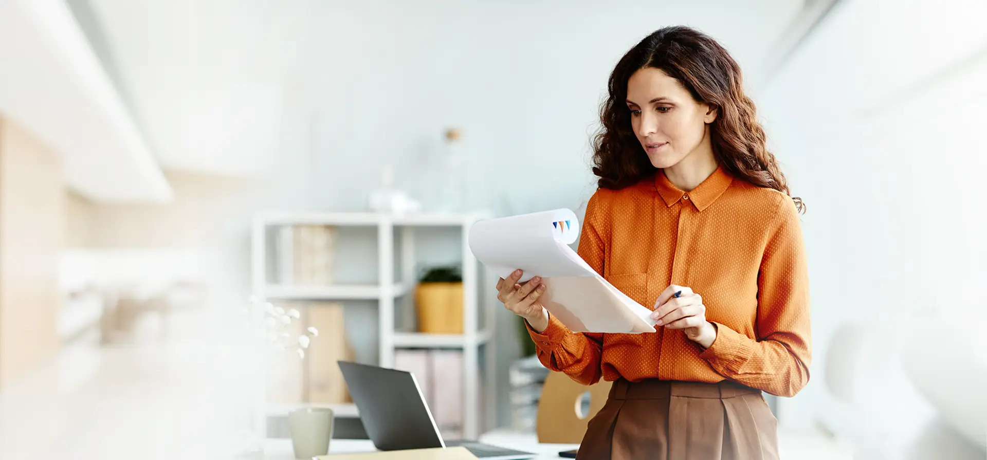 Kobieta w pomarańczowej koszuli analizuje dokumenty z kolorowymi wykresami, trzymając długopis w dłoni, w jasnym biurze z laptopem i regałem w tle.