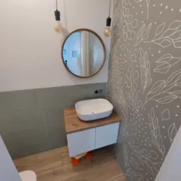 Nowoczesna łazienka z umywalką nablatową na szafce z drewnianym blatem, okrągłe lustro w drewnianej ramie i dekoracyjna tapeta w roślinne wzory na szarej ścianie.