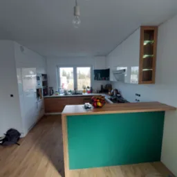 Nowoczesna kuchnia z białymi szafkami, drewnianymi blatami i zielonym barkiem, widok na okno z widokiem na ogród, na blacie kosz z owocami, lampa wisząca na kablu.