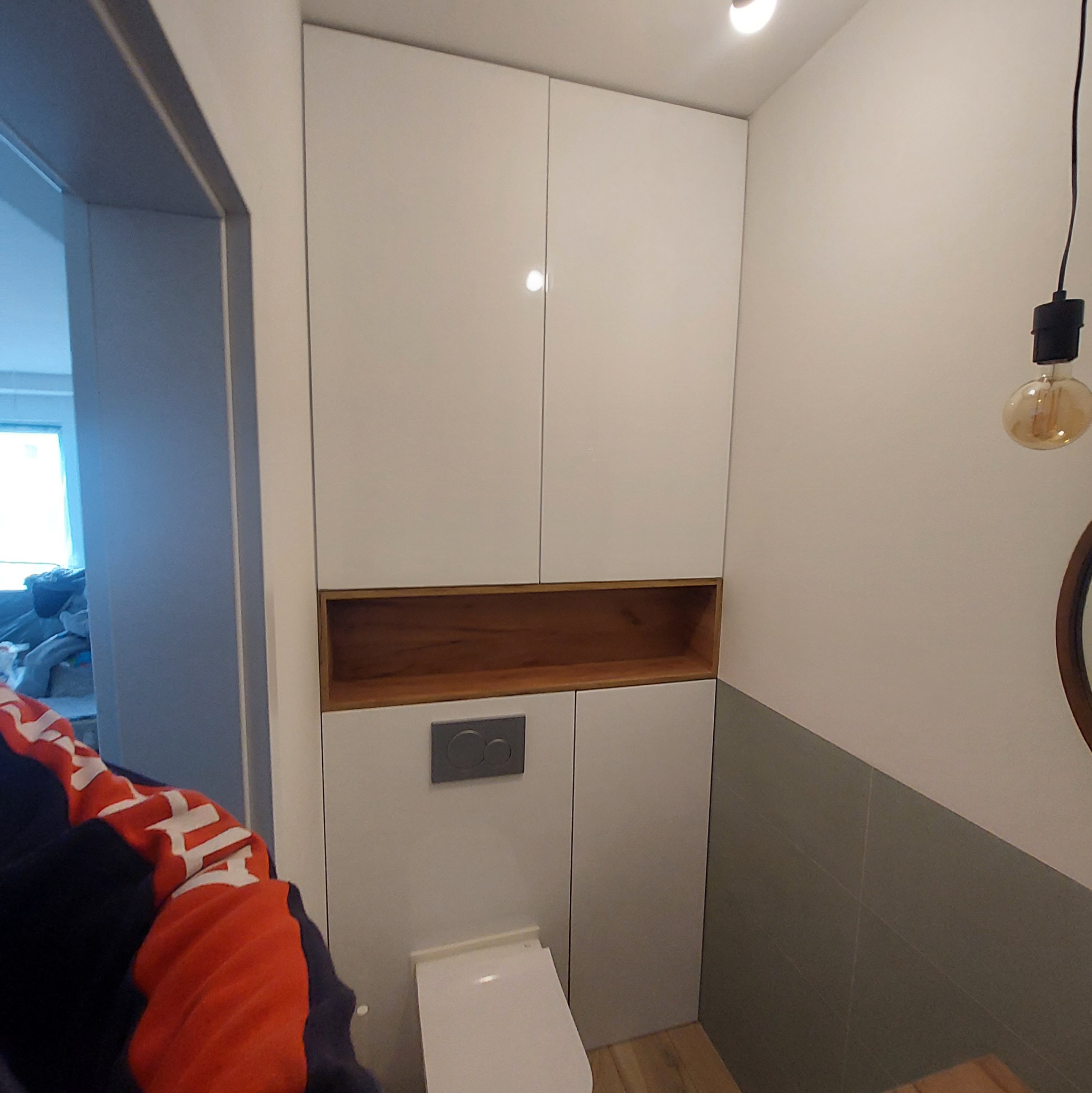 Biała zabudowa w toalecie z drewnianą półką nad spłuczką, widoczny fragment muszli klozetowej i szare płytki na ścianie.