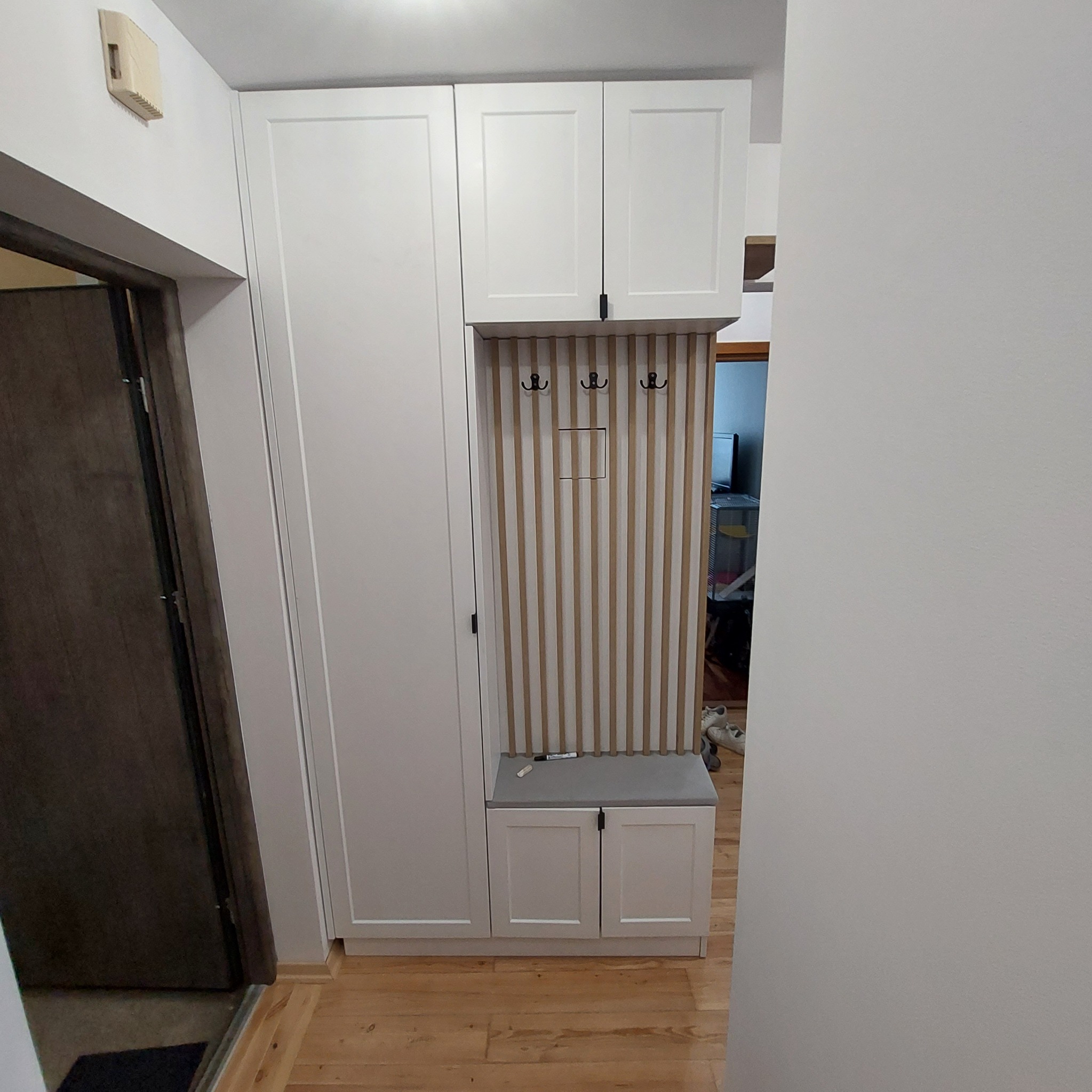 Biała szafa wnękowa z siedziskiem i panelem w pionowe paski, zamontowana w przedpokoju przy wejściu do mieszkania.