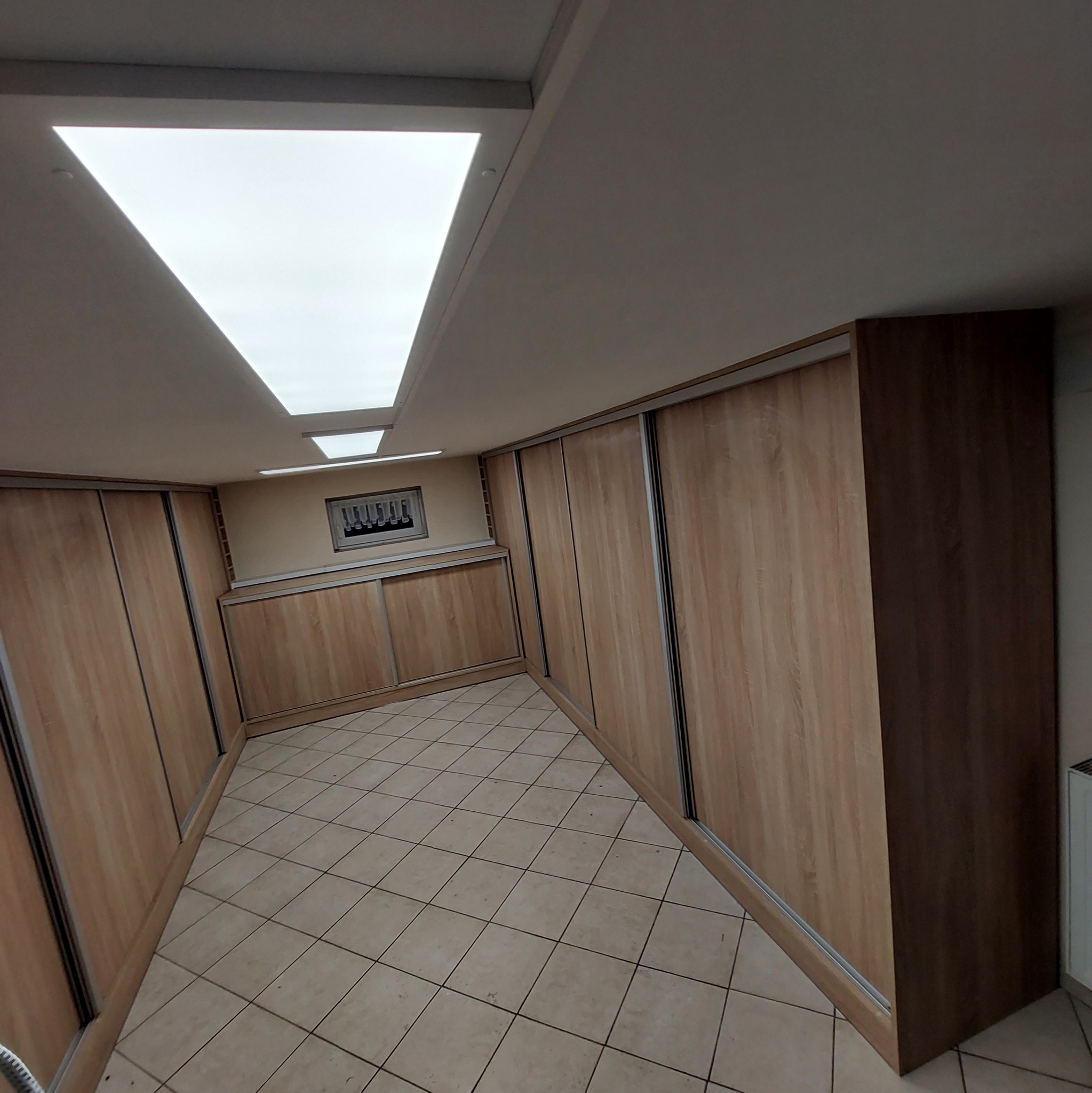 Wnętrze garderoby z zabudowanymi szafami przesuwnymi o jasnym drewnianym wykończeniu, oświetlone panelami sufitowymi, widok z perspektywy podłogi z beżowymi płytkami.