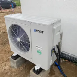 Zewnętrzna jednostka klimatyzacji York zamontowana na betonowych bloczkach na placu budowy, widoczne kable i rury, biała ściana budynku w tle.