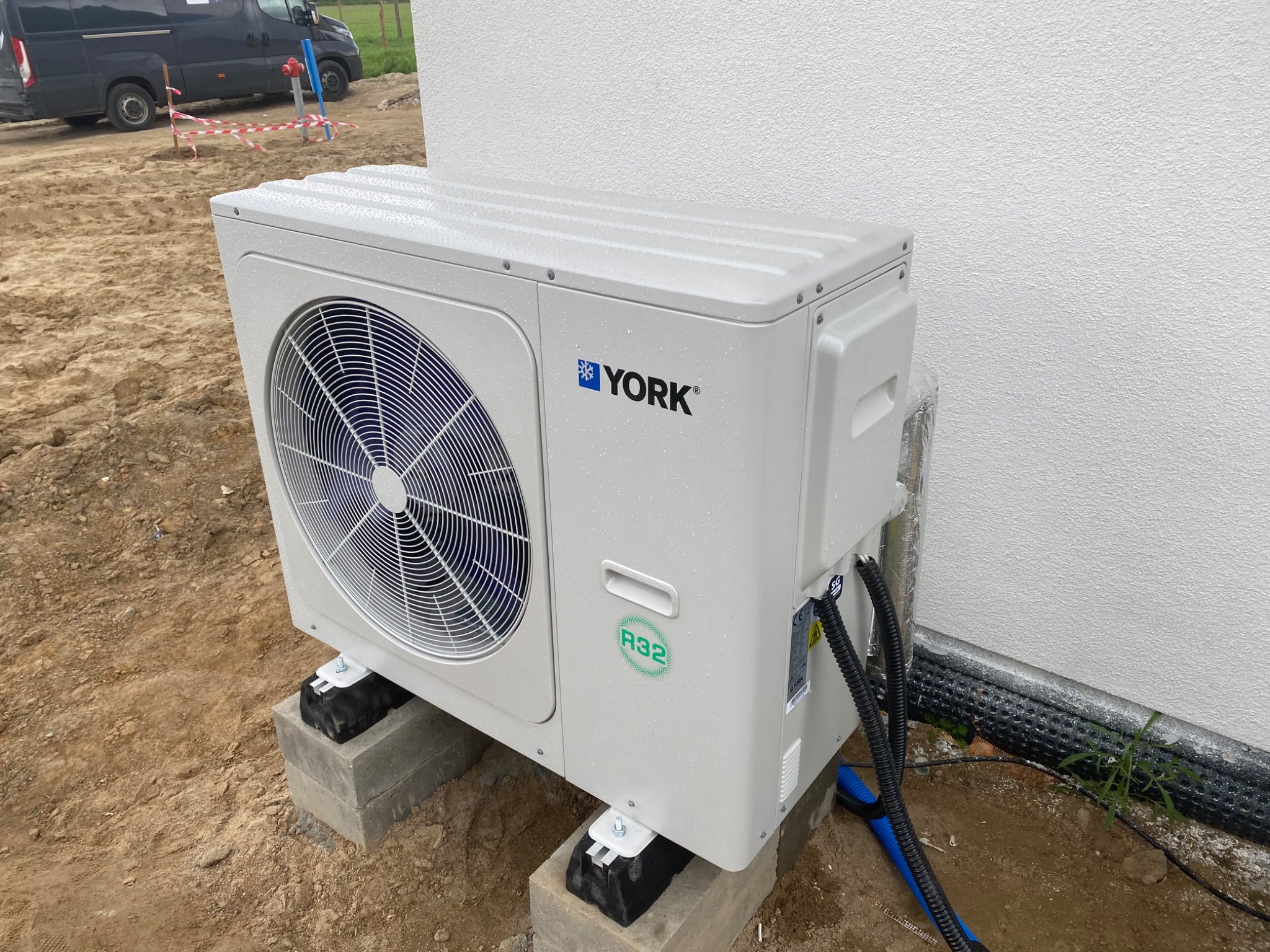 Zewnętrzna jednostka klimatyzacji York zamontowana na betonowych bloczkach na placu budowy, widoczne kable i rury, biała ściana budynku w tle.