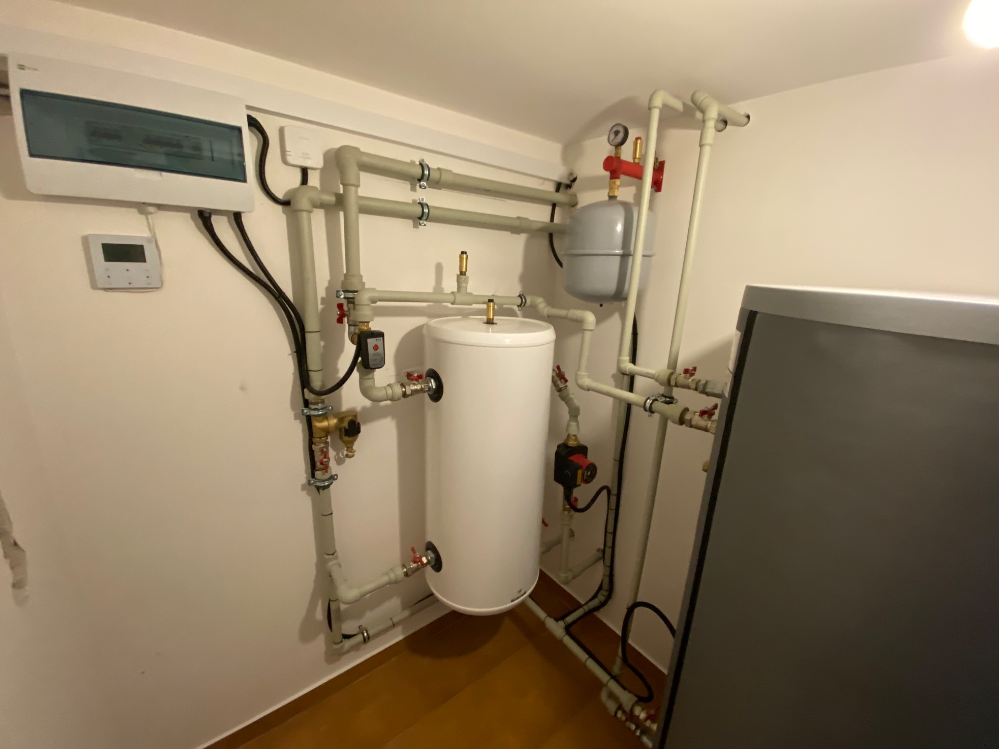 Biały zasobnik ciepłej wody użytkowej z licznymi rurami i zaworami, zamontowany w pomieszczeniu technicznym, widoczne również elementy elektryczne i sterujące.
