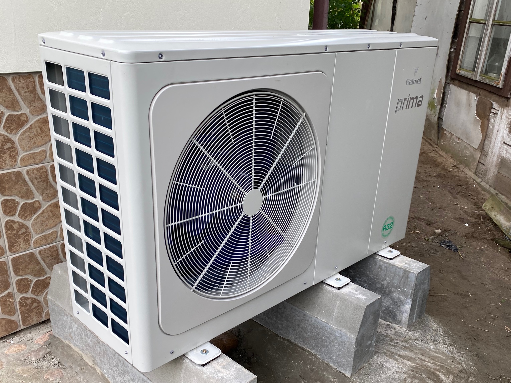 Zewnętrzna jednostka klimatyzacji marki Galmet Prima, zamontowana na betonowych bloczkach przy ścianie z okładziną imitującą kamień, z widocznym symbolem czynnika chłodniczego R32.