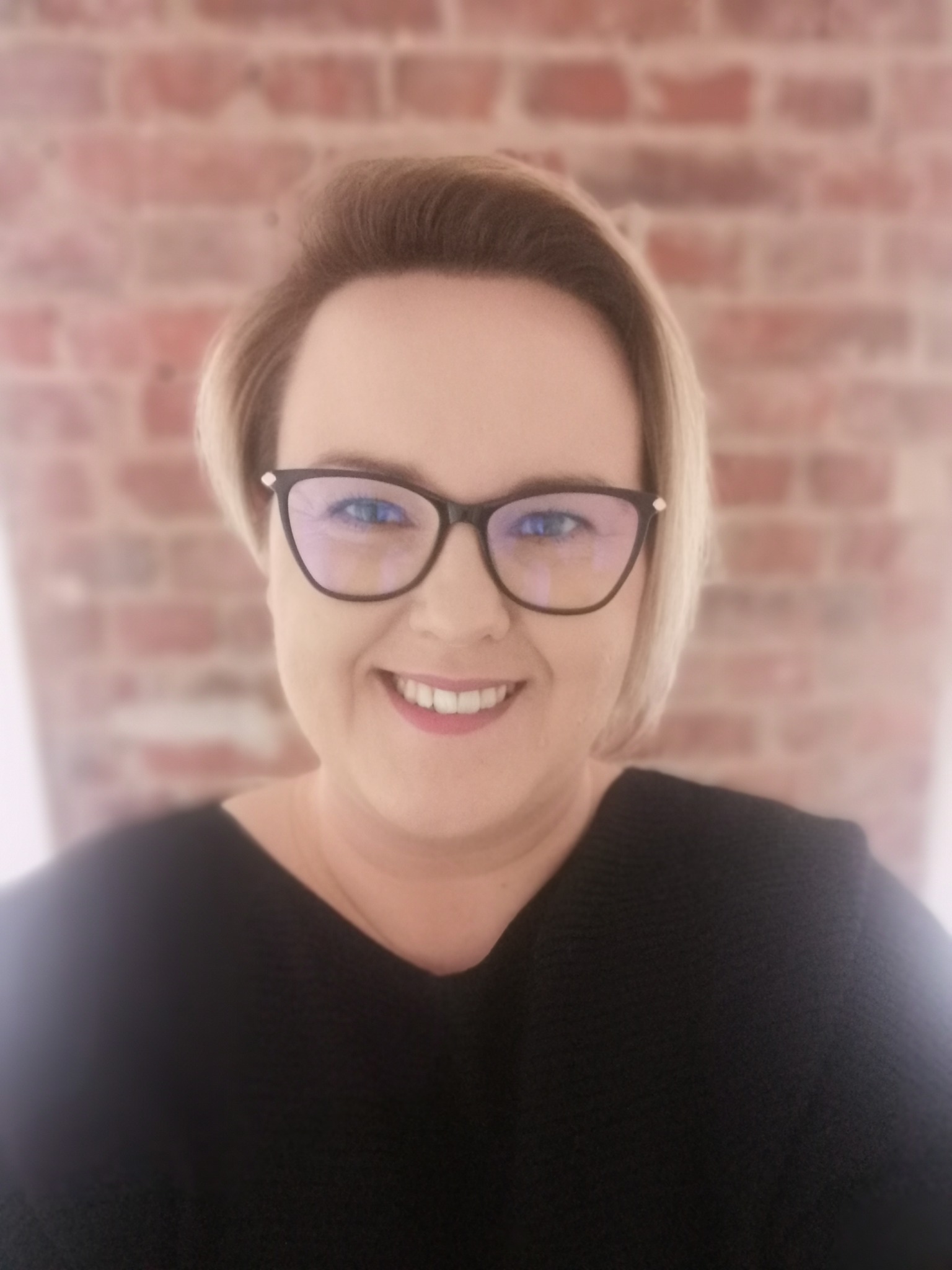 Uśmiechnięta kobieta w okularach, z krótkimi blond włosami, na rozmytym tle ceglanej ściany.