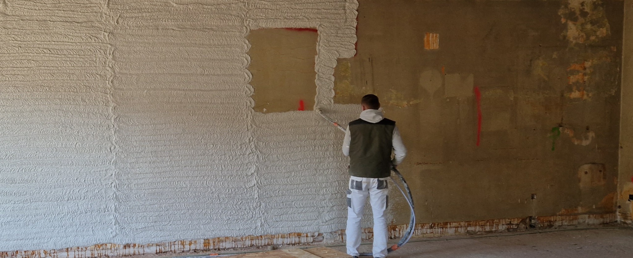 Pracownik w odzieży roboczej nanosi tynk maszynowy o wyraźnej, poziomej fakturze na ścianę w surowym wnętrzu, widoczny fragment ściany bez tynku i ślady po poprzednich pracach.