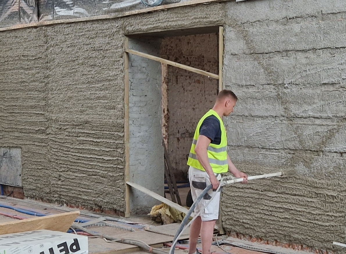 Pracownik w kamizelce odblaskowej nanosi tynk maszynowy na ścianę z widoczną fakturą, w tle surowy mur z cegły w otworze drzwiowym.