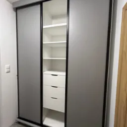 Szafa wnękowa z przesuwnymi drzwiami w kolorze szarym, z czarnymi obramowaniami, wnętrze białe z półkami i trzema szufladami, widok z lekkiego ukosa.