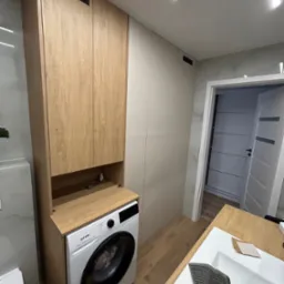 Nowoczesna łazienka z zabudową meblową w odcieniach drewna i bieli, z pralką wkomponowaną w szafkę i wysoką szafą do przechowywania, widok na otwarte drzwi do kolejnego pomieszczenia.