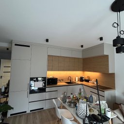 Stelmont Filip Stelmach - Nowoczesna kuchnia z szarymi i drewnianymi frontami, oświetleniem LED pod szafkami wiszącymi i czarnymi akcentami, z widocznym bałaganem na stole jadalnym.