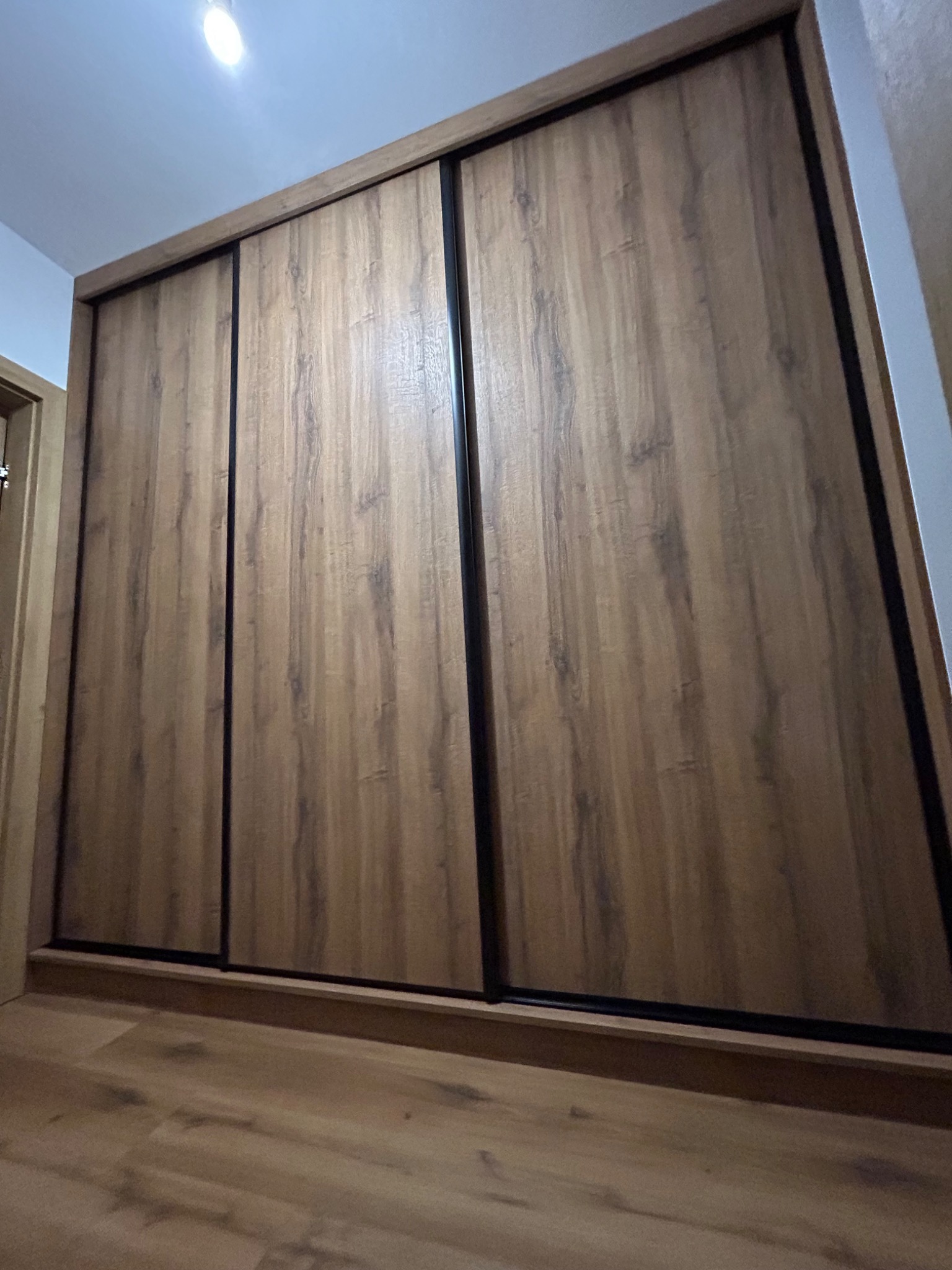 Trzydrzwiowa szafa przesuwna z okładziną imitującą drewno, z czarnymi profilami, widoczna z dołu, na tle podłogi w podobnym odcieniu.