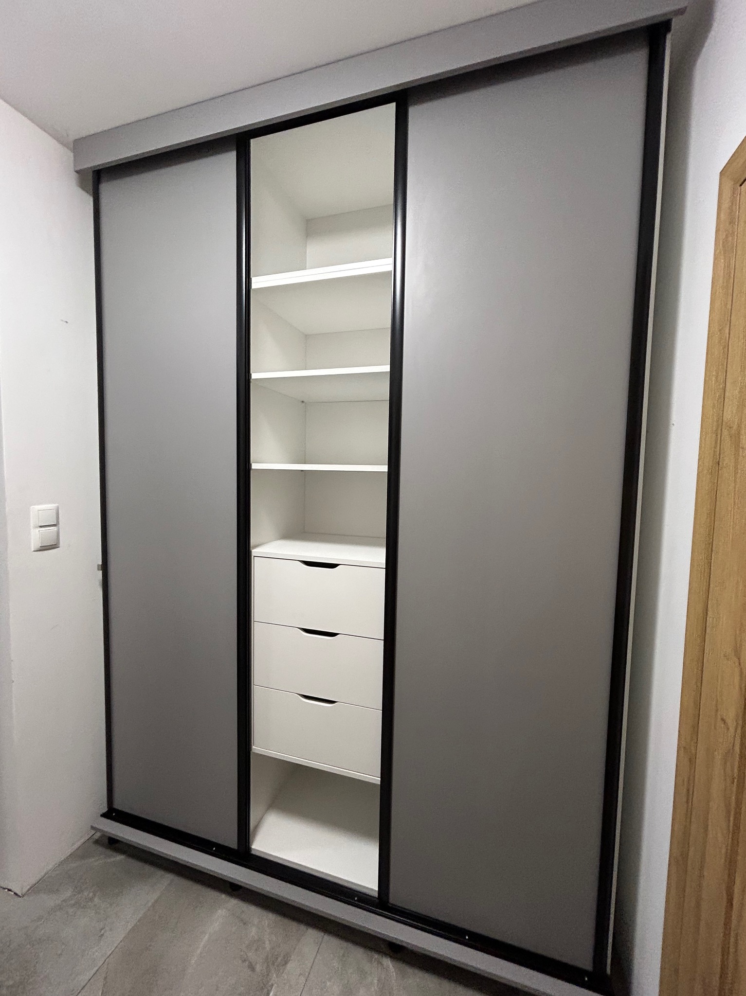 Szafa wnękowa z przesuwnymi drzwiami w kolorze szarym, z czarnymi obramowaniami, wnętrze białe z półkami i trzema szufladami, widok z lekkiego ukosa.