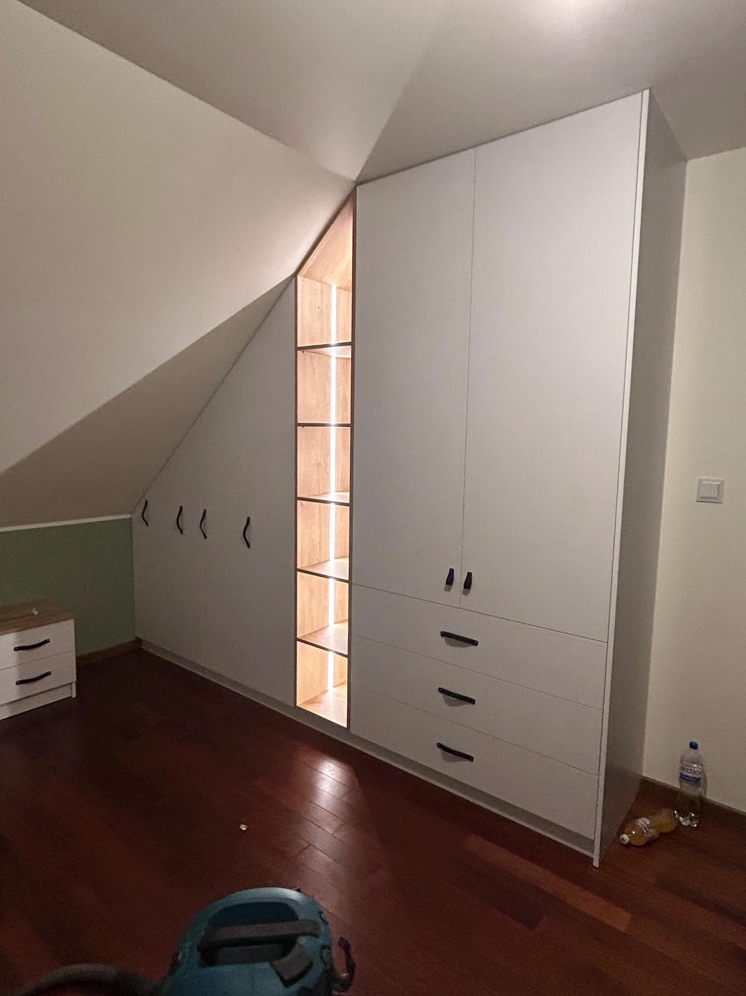 Biała szafa z szufladami i otwartą częścią z półkami podświetlonymi od wewnątrz, dopasowana do skosu poddasza, widoczne wieszaki na ścianie, na podłodze butelki i narzędzie.