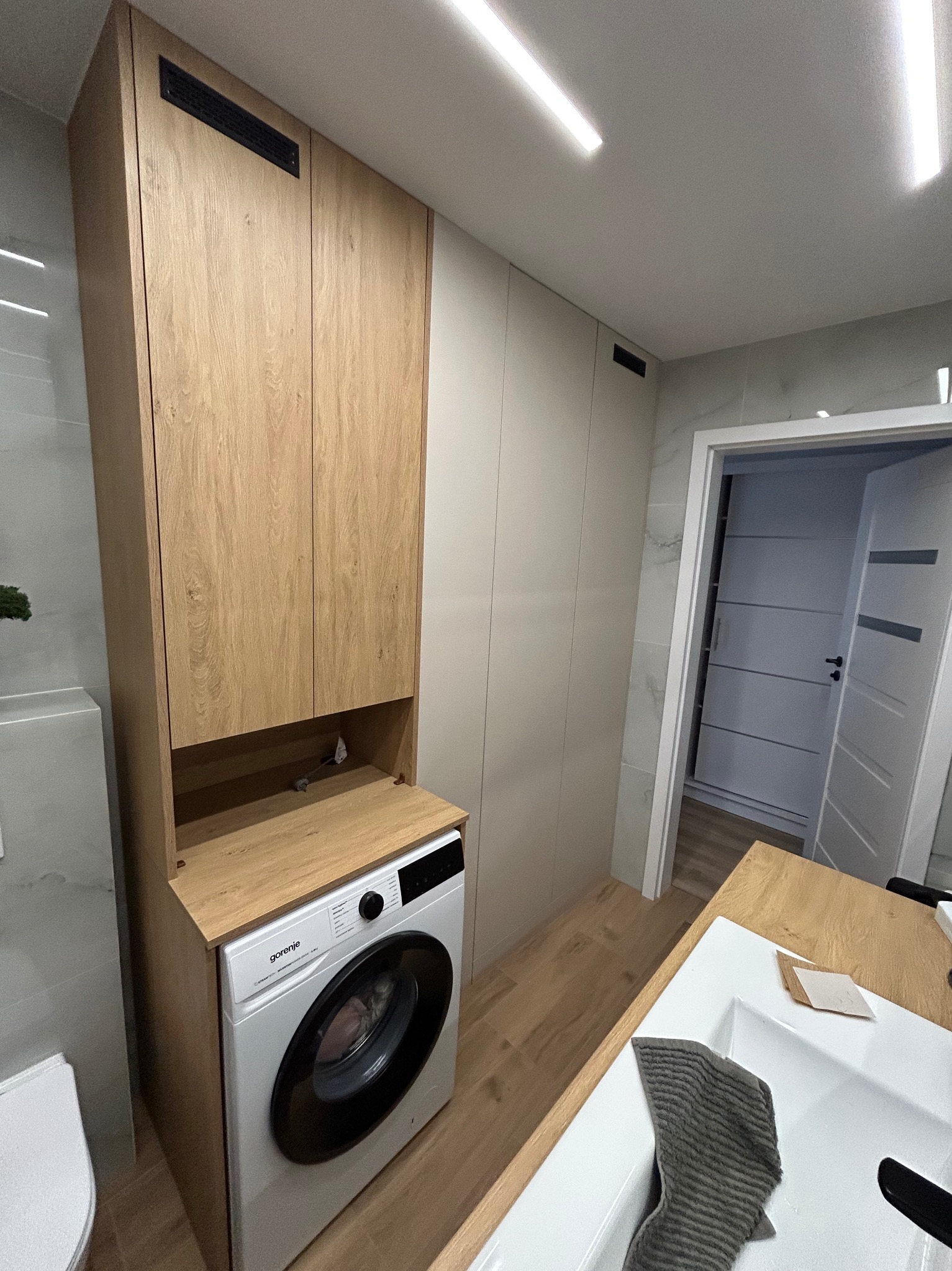 Nowoczesna łazienka z zabudową meblową w odcieniach drewna i bieli, z pralką wkomponowaną w szafkę i wysoką szafą do przechowywania, widok na otwarte drzwi do kolejnego pomieszczenia.