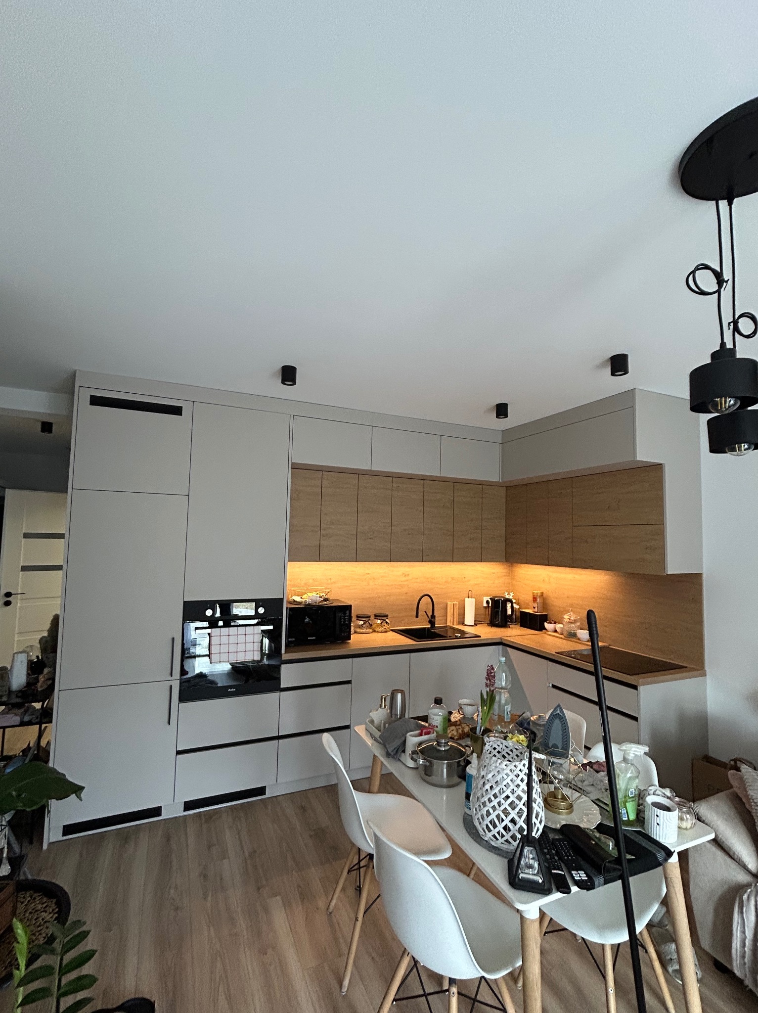 Nowoczesna kuchnia z szarymi i drewnianymi frontami, oświetleniem LED pod szafkami wiszącymi i czarnymi akcentami, z widocznym bałaganem na stole jadalnym.