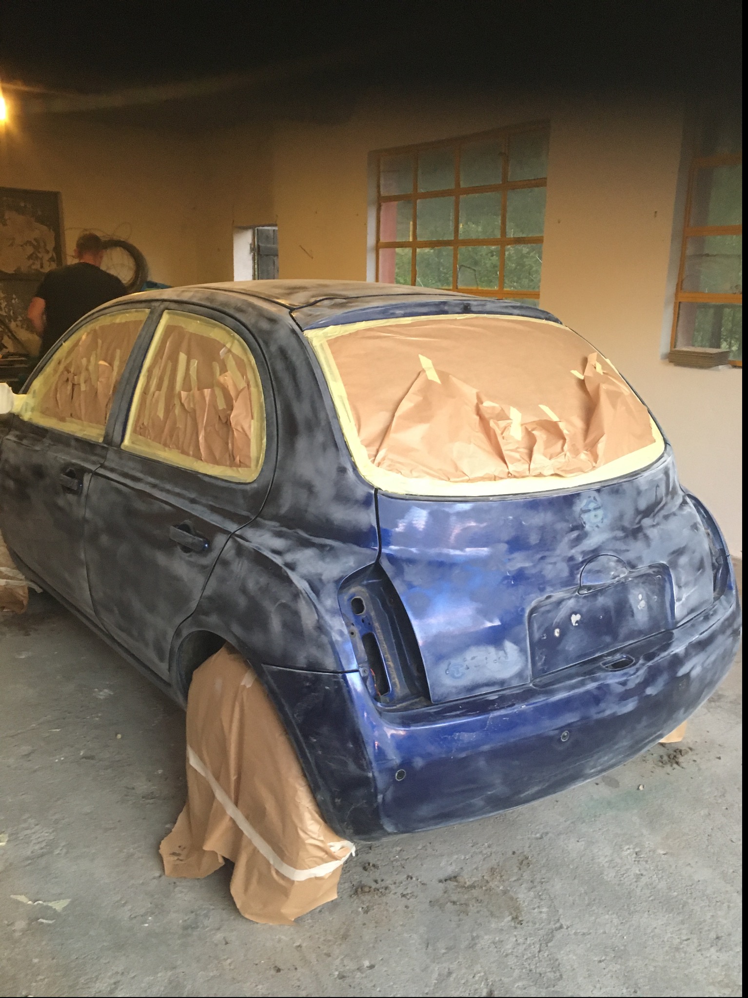 Przygotowanie niebieskiego samochodu do lakierowania: oklejone szyby i elementy karoserii papierem i taśmą maskującą, widoczne ślady szpachlowania, w tle warsztat z oknami i pracownikiem.