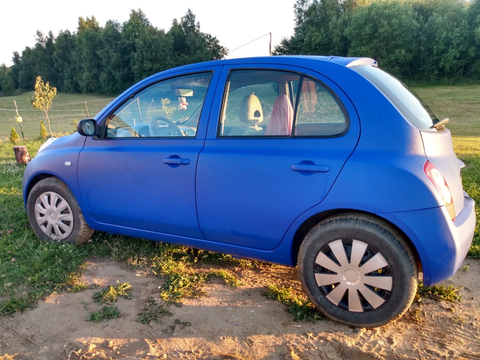 Niebieski Nissan Micra po lakierowaniu z fakturą, stojący na trawie, widok z boku, zarys lasu w tle.