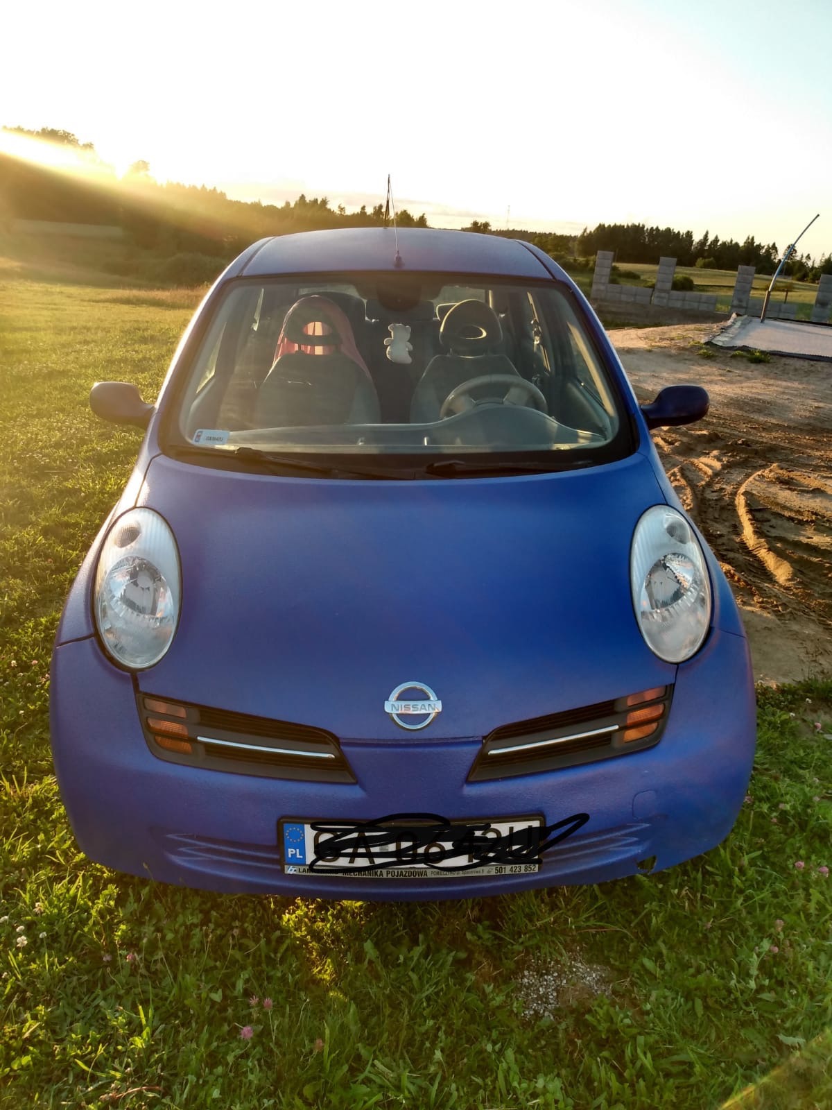 Niebieski Nissan Micra po amatorskim lakierowaniu, widoczne niedoskonałości powłoki, zaparkowany na trawie z widokiem na zachodzące słońce.