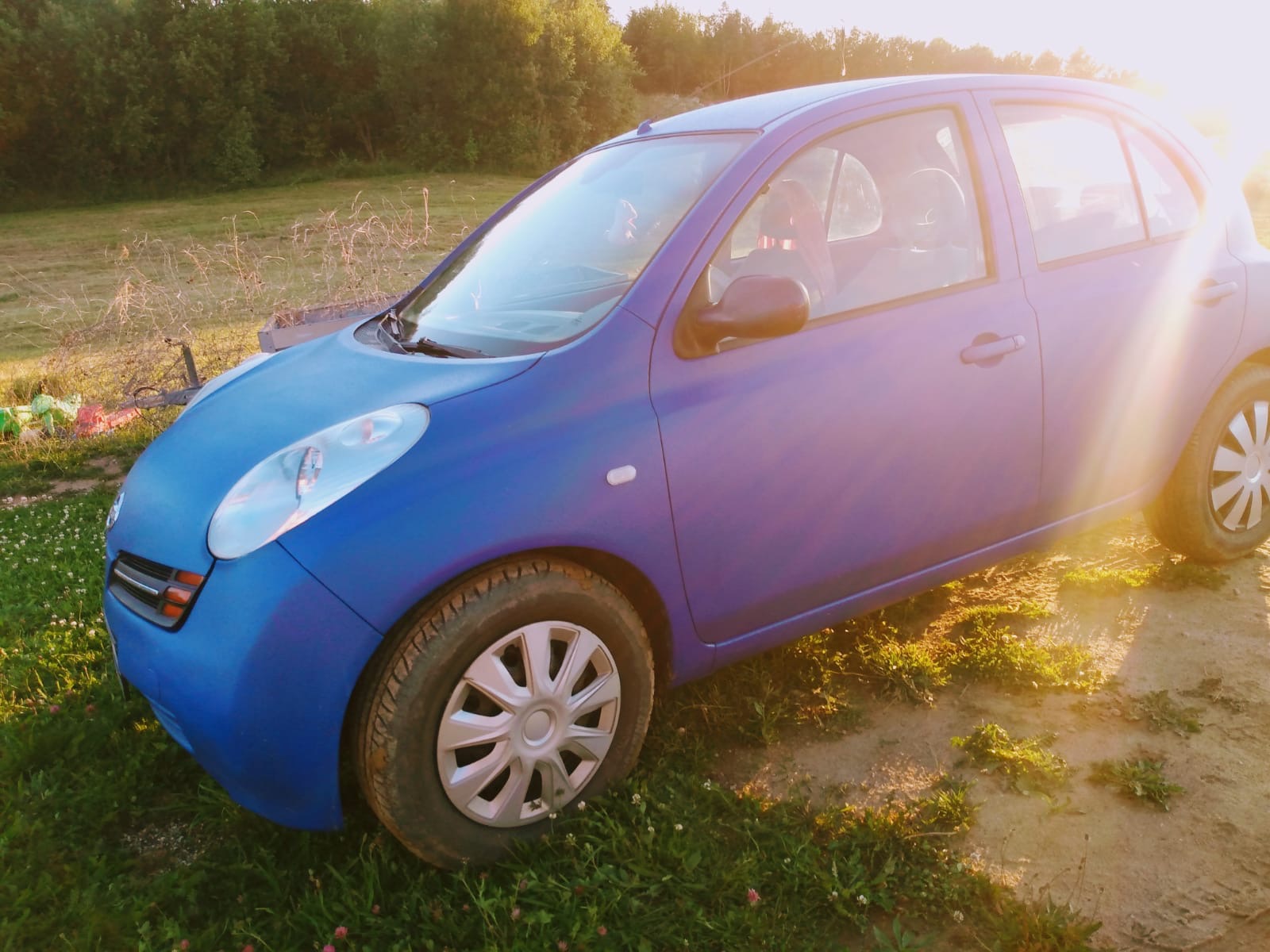 Niebieski samochód osobowy Nissan Micra po lakierowaniu, stojący na trawiastym terenie w promieniach zachodzącego słońca.