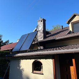 GreenTech - Dwa panele słoneczne zamontowane na dachu budynku jednorodzinnego z ceglanym kominem i anteną satelitarną, błękitne niebo w tle.