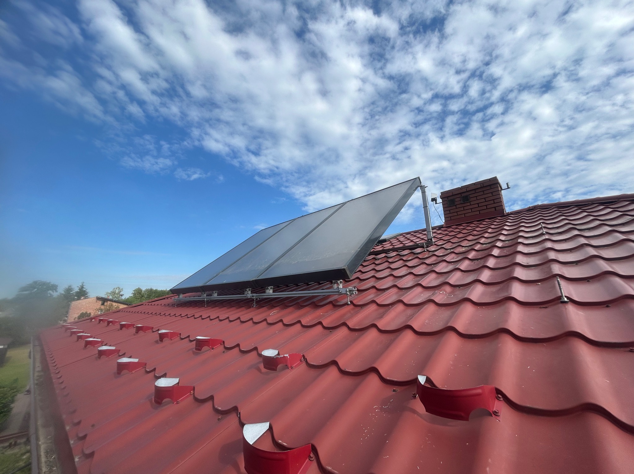 Panel słoneczny zamontowany na czerwonym dachu z dachówki ceramicznej, widok z dołu na błękitne niebo z chmurami.
