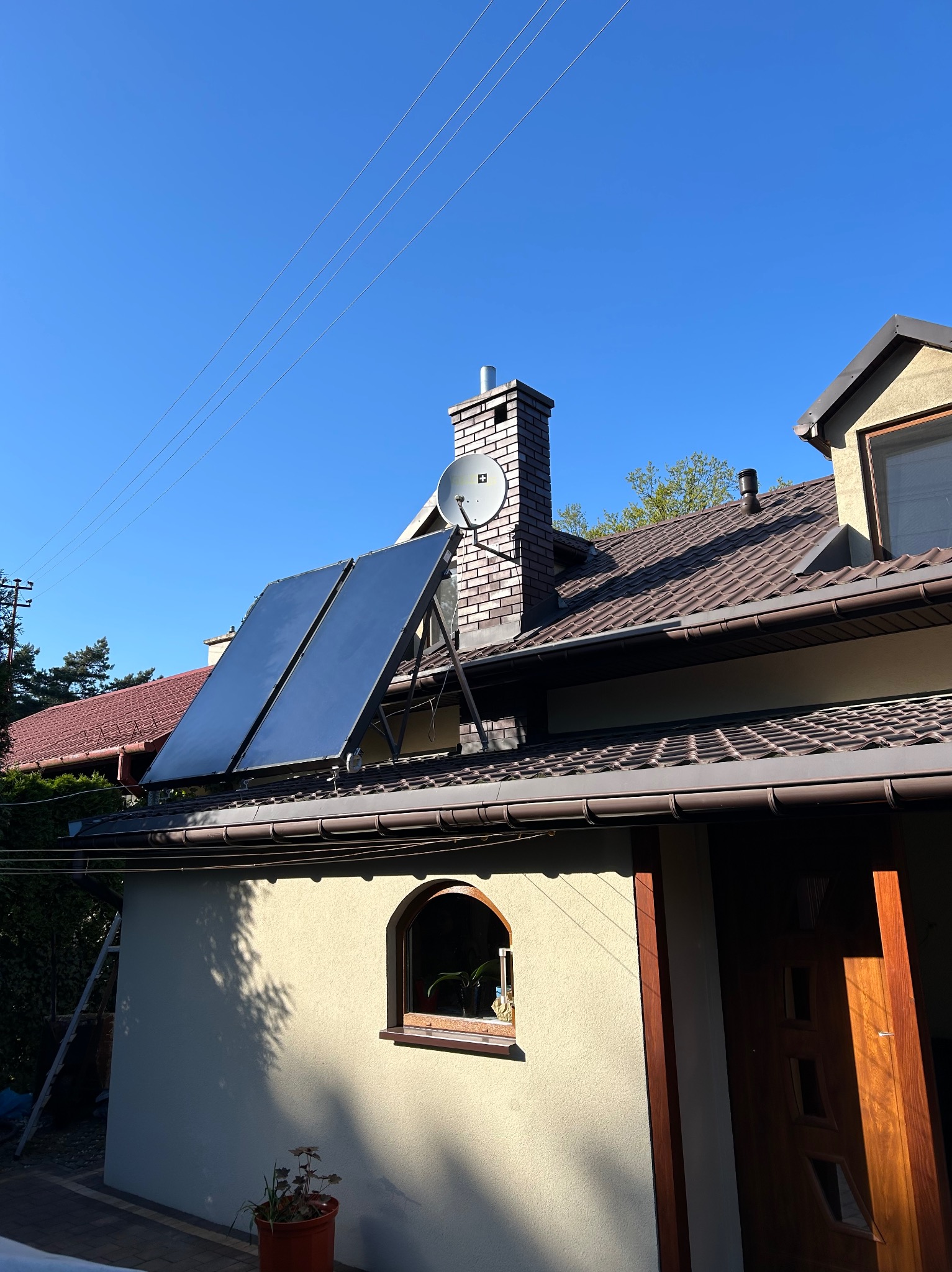 Dwa panele słoneczne zamontowane na dachu budynku jednorodzinnego z ceglanym kominem i anteną satelitarną, błękitne niebo w tle.