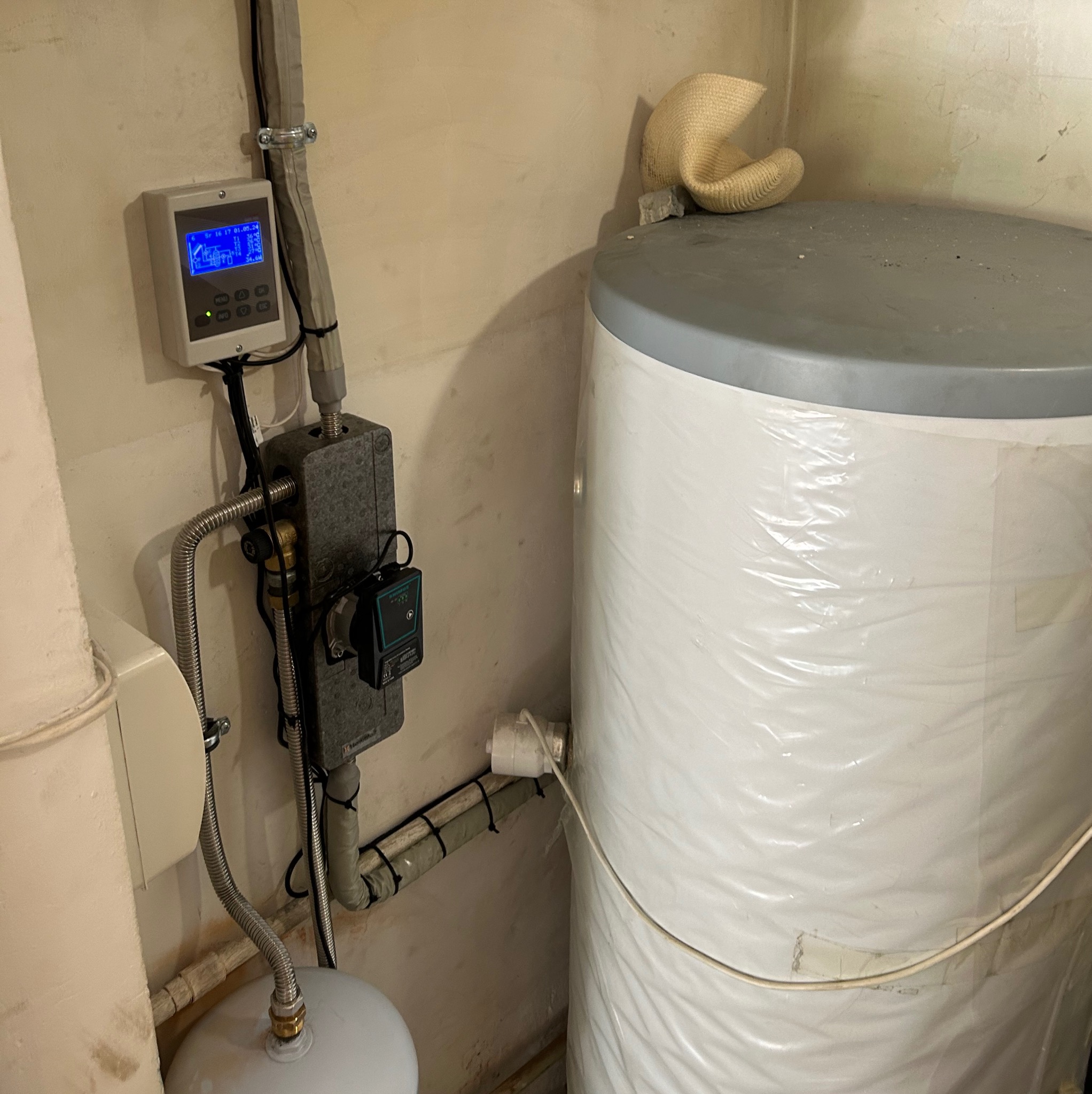 Bojler owinięty folią, z podłączoną instalacją hydrauliczną, zaworem i sterownikiem elektronicznym na ścianie w surowym pomieszczeniu.