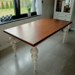 Rozkładany drewniany stół z blatem w odcieniach brązu i toczonymi, kremowymi nogami na tle okna z widokiem na ogród i białej witryny.