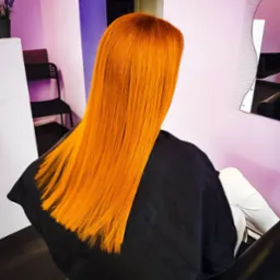 Klientka z długimi, prostymi, intensywnie pomarańczowymi włosami siedzi w fotelu fryzjerskim, ubrana w czarną pelerynę ochronną, widok z tyłu.