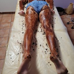 ALEKSANDRA KONOPNICKA - Osoba leżąca na stole zabiegowym, pokryta brązową maską na ciele, w jednorazowej bieliźnie i czepku.