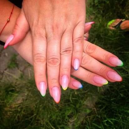 Dłonie z pomalowanymi paznokciami w odcieniach różu i z kolorowymi końcówkami (żółty, niebieski, zielony, czerwony) na tle trawy.