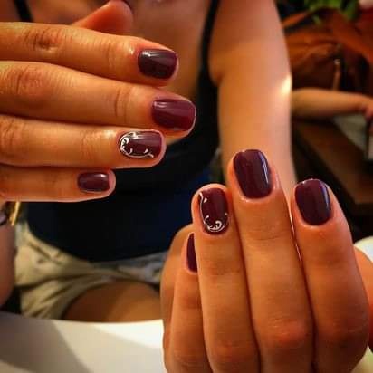 Dłonie z ciemnoczerwonym lakierem na paznokciach, zdobienia w postaci srebrnych wzorów na dwóch paznokciach.