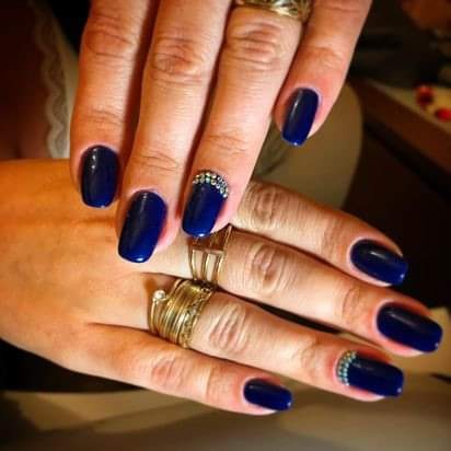 Dłonie z granatowym manicure hybrydowym, zdobionym rzędem drobnych kryształków przy skórkach, oraz złotymi pierścionkami na palcach.