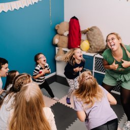 RATATATA SP. Z O.O. - Animator z grupą dzieci podczas zabawy w sali z niebieską ścianą i dekoracjami. Wszyscy pokazują języki, wyrażając radość i zaangażowanie w aktywność.