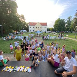 RATATATA SP. Z O.O. - Plenerowa impreza dla dzieci z animatorem na scenie, w tle zabytkowy budynek i publiczność. Dzieci trzymają żółte rekwizyty, animator z mikrofonem.