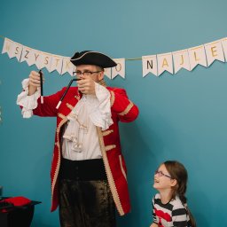 RATATATA SP. Z O.O. - Iluzjonista w stroju pirata prezentuje trik z różdżkami, a dziewczynka w okularach z radością obserwuje pokaz na tle błękitnej ściany z napisem 'Wszystkiego Najlepszego'.